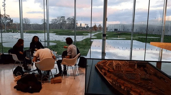 Quatre personnes sont assises sur des chaises blanches autour d'une table dans un bâtiment moderne aux murs de verre avec une vue sur l'herbe à l'extérieur pendant le Culturathon 2018. Des ordinateurs portables, des sacs et du matériel sont posés sur le sol ; une maquette d'architecture en bois est posée sur un support au premier plan.