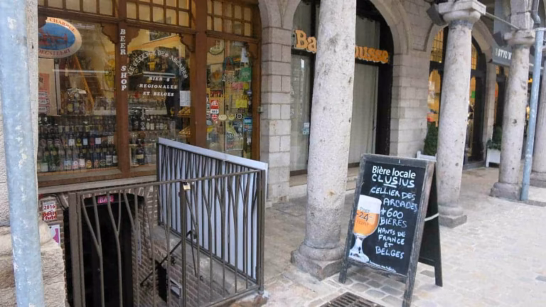 Une arcade en pierre avec des boutiques - parfaites pour faire du shopping à Arras - comporte un panneau à craie annonçant de la bière locale. Une rampe en métal entoure les escaliers menant à un magasin de bière, visible à travers une vitre avec des bouteilles à l'intérieur et des autocollants colorés sur la porte. Rue pavée au premier plan.