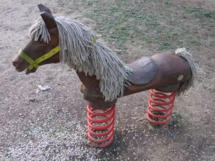 Un cheval à bascule en bois avec un harnais jaune, une crinière et une queue en laine, et des ressorts métalliques rouges en guise de pattes, se dresse sur une pelouse inégale - parfait pour une sortie en famille à Arras, montrant de charmants signes d'utilisation.